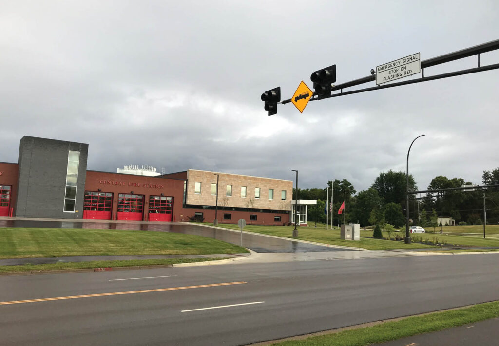 A street view showcases the Central Fire Station, featuring red garage doors and a neatly mowed lawn. Above the street, a traffic light and road sign symbolize efforts in enhancing emergency vehicle access to public roadways for quicker response times.