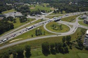Aerial view of a highway interchange with multiple looping ramps. Vehicles are lined up, causing traffic congestion on one side. Surrounding areas include green spaces, trees, a parking lot, and open fields.