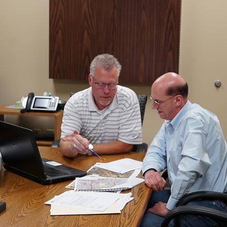 Two men are seated at a wooden table in an office, looking at papers and a laptop. One man, wearing glasses and a striped polo shirt, is pointing at the documents. The other man, in a light blue shirt, is attentively listening.