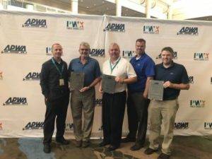 Five men standing in front of a backdrop with APWA and PWX Public Works Expo logos, holding plaques. They are smiling and dressed in business casual attire. The setting appears to be an indoor event or conference.