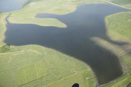 Aerial view of a lake shaped like a dinosaur with outstretched arms on grassland. The lakes dark water contrasts with the light green landscape surrounding it. Crops and fields are visible in the foreground.