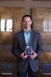 A person in a blue shirt and gray blazer is holding an award. They are standing in front of a textured, gold-colored wall with square art pieces.
