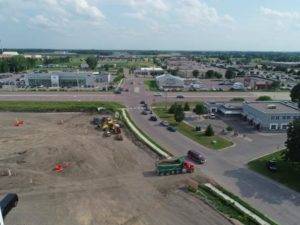 Aerial view of a construction site near a road intersection. Cars are visible on the roads beside the site. Nearby, various buildings and greenery surround the area under a partly cloudy sky.