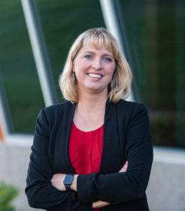 A woman with blonde hair in a black blazer and red blouse, representing the MN Young American Leaders Program, smiles confidently with arms crossed. She stands poised in front of a building with large windows.