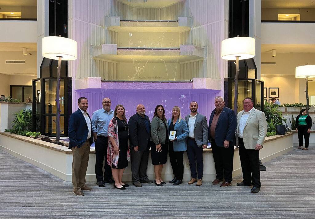 A group of ten people stands in front of a large, illuminated waterfall feature inside a building. They are dressed in formal attire and are smiling at the camera. The setting includes plants and modern lighting fixtures.