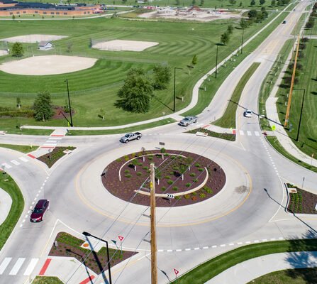 Aerial view of a traffic roundabout surrounded by green grass and landscaped areas. Nearby, a baseball field and other open spaces are visible. Cars are navigating the roundabout, and streets lead in multiple directions.