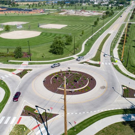 Aerial view of a traffic roundabout surrounded by green grass and landscaped areas. Nearby, a baseball field and other open spaces are visible. Cars are navigating the roundabout, and streets lead in multiple directions.