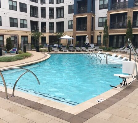 A modern apartment complex surrounds a rectangular outdoor pool with clear blue water. Lounge chairs and umbrellas line the poolside, and greenery decorates the area. The building features a mix of white and brick facade.