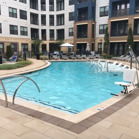 A modern apartment complex surrounds a rectangular outdoor pool with clear blue water. Lounge chairs and umbrellas line the poolside, and greenery decorates the area. The building features a mix of white and brick facade.