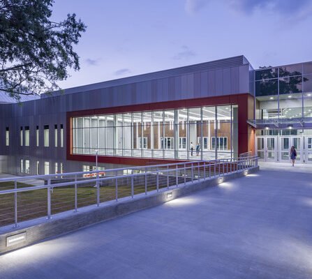 Modern building with large glass windows and a prominent red frame around a central section. A walkway with metal railings leads to the entrance. The scene is lit with soft evening lighting, and a tree is visible on the left.