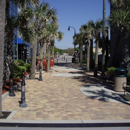 A walkway lined with tall palm trees leads to a street. Decorative red urns are placed along the path. Buildings with glass windows are on the left, and street lamps line the pathway. Bright sunlight casts shadows on the pavement.