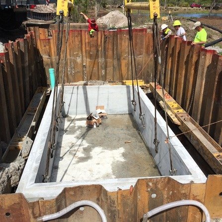 A large rectangular concrete pit in a construction site, secured with metal beams and surrounded by wooden barriers. Several workers in safety gear are visible in the background.