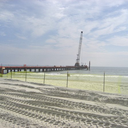 A sandy beach with tire tracks leading to a long pier extending into the ocean. A crane is positioned on the pier. The sky is partly cloudy, and the water appears calm. A fence runs along the beach, parallel to the shoreline.