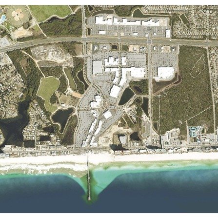 Aerial view of a coastal town showing a beachfront with a pier extending into the ocean. There are buildings, parking lots, and green spaces behind the beach. Roads and residential areas are visible further inland.