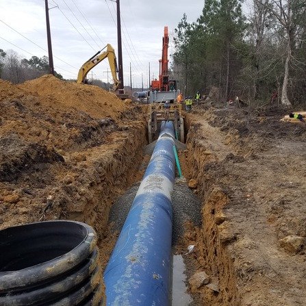 Construction site with workers installing a large blue pipe in a trench. Excavators are visible, and surrounding areas have piles of dirt and forested land. Power lines run parallel to the trench.