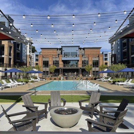 A modern outdoor pool area at an apartment complex. The scene includes a central pool, surrounded by lounge chairs with blue umbrellas, a round fire pit with wooden chairs, and string lights overhead. A multi-story building is in the background.