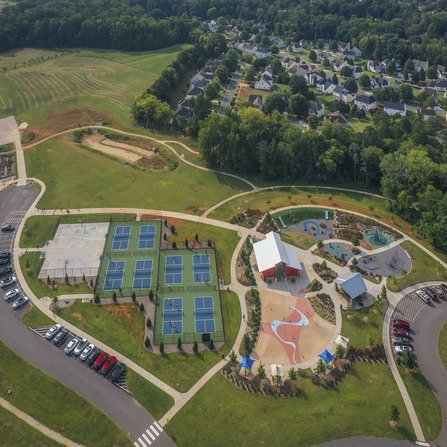 Aerial view of a recreational park with tennis courts, a playground, and a splash pad. A parking lot is on the left, and a residential neighborhood and forested area are in the background. Pathways connect the different areas of the park.