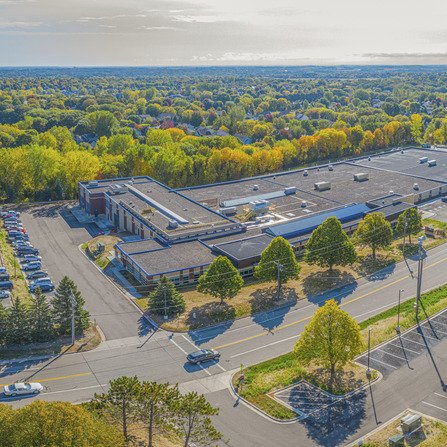 Aerial view of a spacious building and car-filled parking lot, surrounded by an abundance of trees