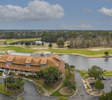 Aerial view of a golf course with a clubhouse in the foreground. The clubhouse has a rust-colored roof and an American flag nearby. The course features lush green fairways, sand traps, and a pond. Trees and a cloudy sky are in the background.