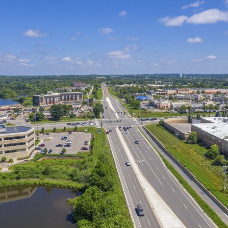 Aerial view of a highway cutting through a mix of residential areas, offices, large buildings, and a lush wooded area
