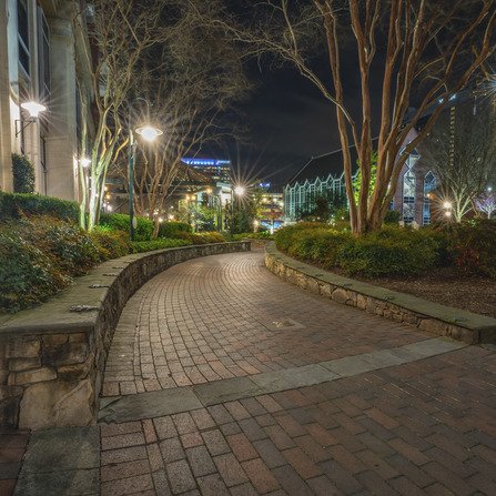 Brick walkway winding through a landscaped area at night, bordered by leafless trees and illuminated by streetlights. Buildings in the background have lights, enhancing the serene urban atmosphere.