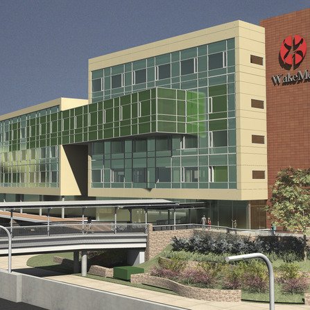 Modern building featuring large glass windows and green accents. The structure has multiple levels, with an overpass in the foreground. There is a logo on one side of the building, and landscaping with greenery around the entrance.