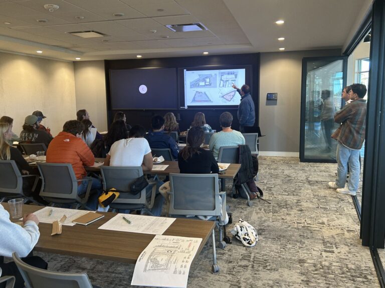 A group of people sits at tables in a classroom, engaged in a presentation on STEM/STEAM careers. The presenter points at architectural designs projected on a screen. Papers and laptops scatter the tables, while another person leans against a glass wall in the background.