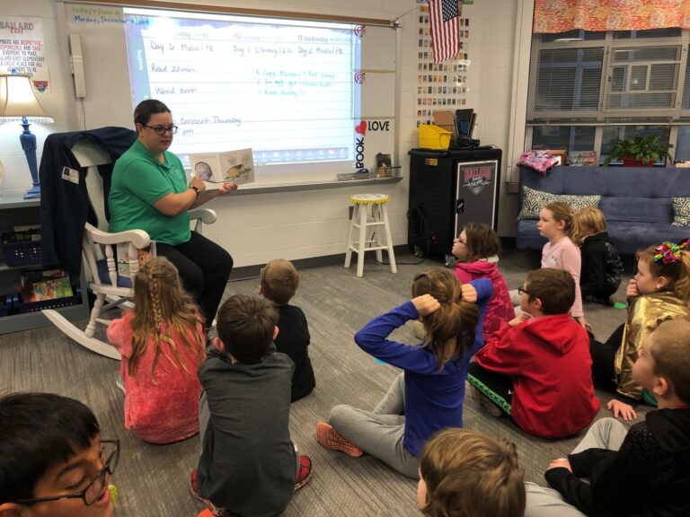A teacher sits in a rocking chair, reading a book to young students seated on the floor. The smart board displays a schedule, hinting at future STEM careers. Children listen attentively in the room filled with educational materials.
