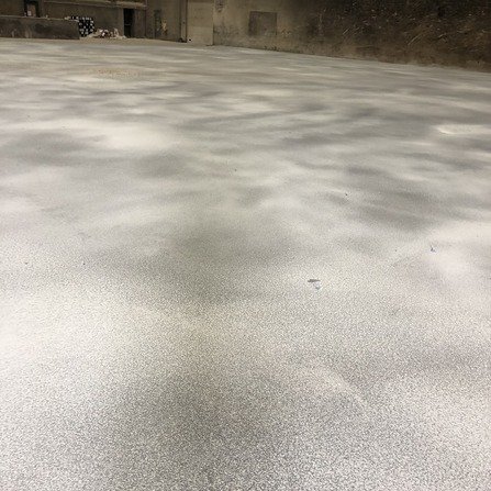 A textured, empty surface with a concrete-like appearance fills the foreground. The background shows a few small, indistinct structures against a blank wall. The overall scene appears industrial and desolate.