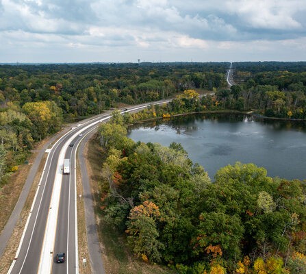 Aerial view of a highway curving alongside a large lake surrounded by dense green forests. Several vehicles are on the road, and the sky is cloudy, casting a soft light over the landscape.