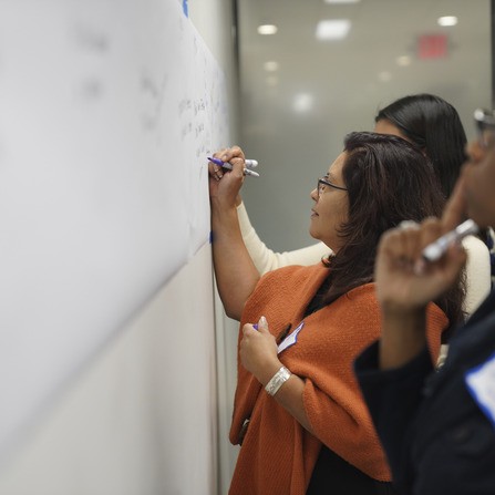 Three people are writing on a large whiteboard. The woman in the foreground is wearing glasses and an orange shawl, writing with a purple marker. The scene suggests a collaborative or educational setting.