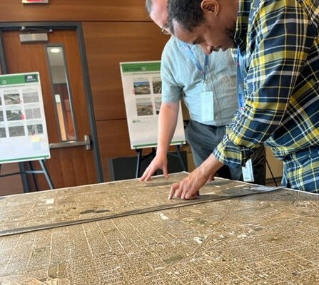 Two people examine a large map on a table in a room with wood-paneled walls. Posters with diagrams are displayed on easels in the background. They are focused, pointing at different sections of the map.