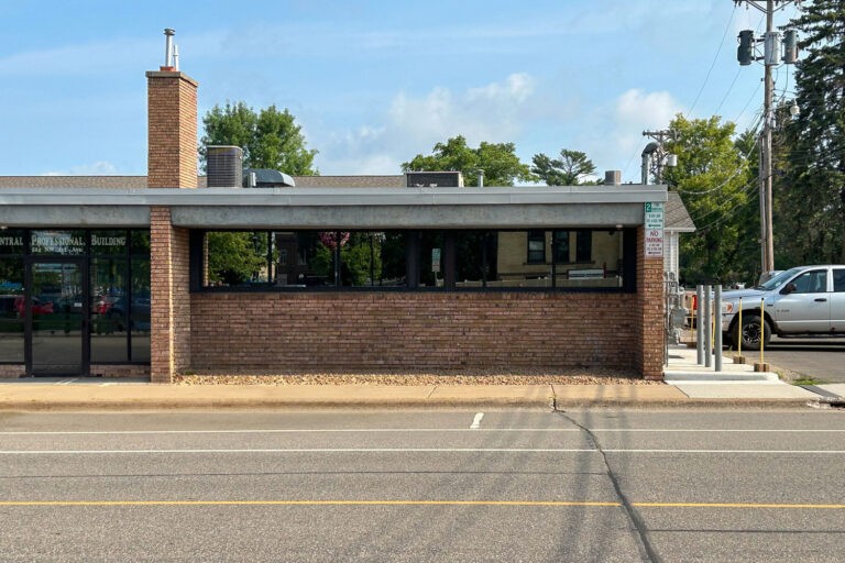 A single-story brick building with a chimney, large windows, and an entrance on the left stands proudly among Bolton & Menk locations. A well-marked parking area is out front, bordered by a road, while trees and utility poles frame the scene in the background.
