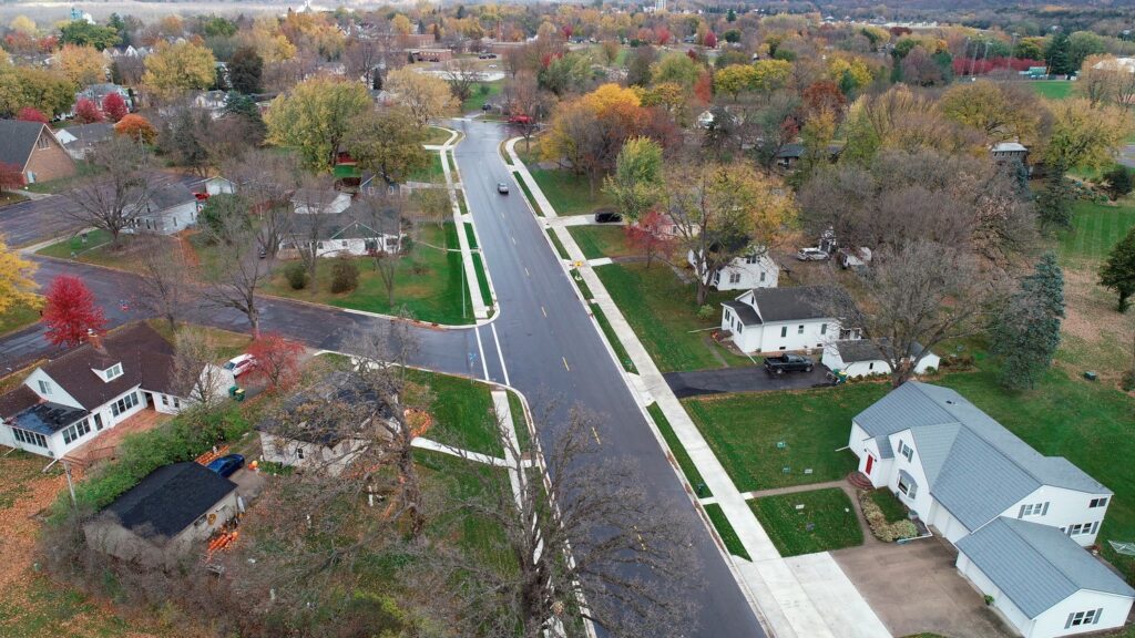 Aerial view of a suburban neighborhood with a wide, empty street perfect for walking or biking, lined by houses and trees. The area displays fall foliage in vibrant shades of red, orange, and yellow. Well-kept lawns extend from the houses, while a distant landscape is visible.