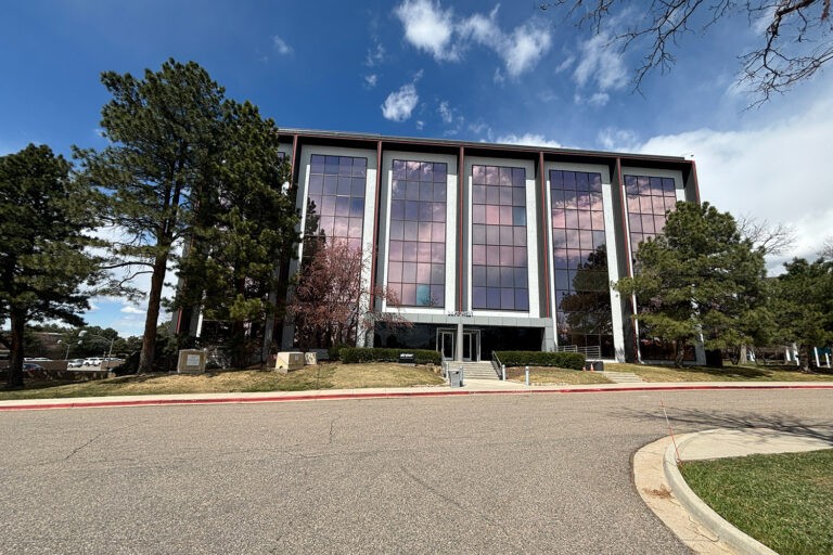 A modern four-story office building with large reflective windows stands among tall pine trees, one of the distinctive Bolton & Menk locations. The partly cloudy sky casts its reflections on the glass as a curved roadway leads to a grassy lawn at the entrance.