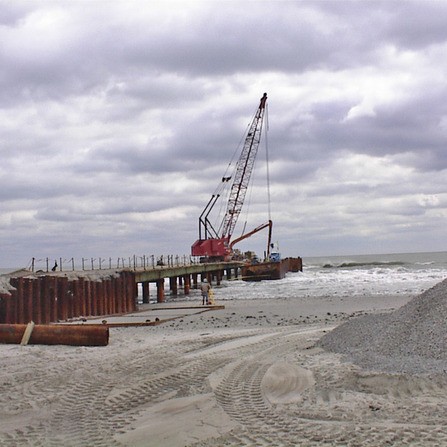 A construction site by the sea with a crane on a pier extending into the water. Steel beams and gravel piles are visible in the foreground, under a cloudy sky. Waves reach the base of the pier.