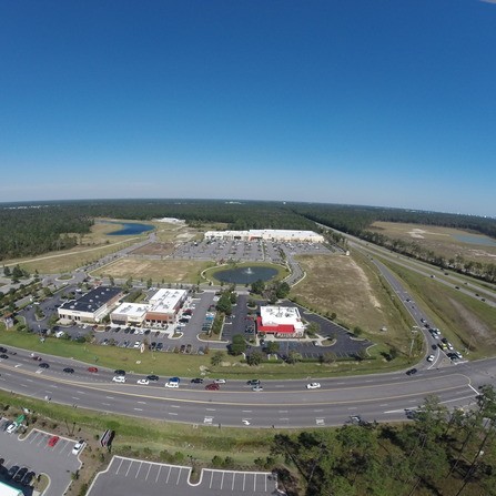 Aerial view of a shopping area with several parking lots, a pond, and a busy road. The background shows a vast expanse of greenery and a clear blue sky.