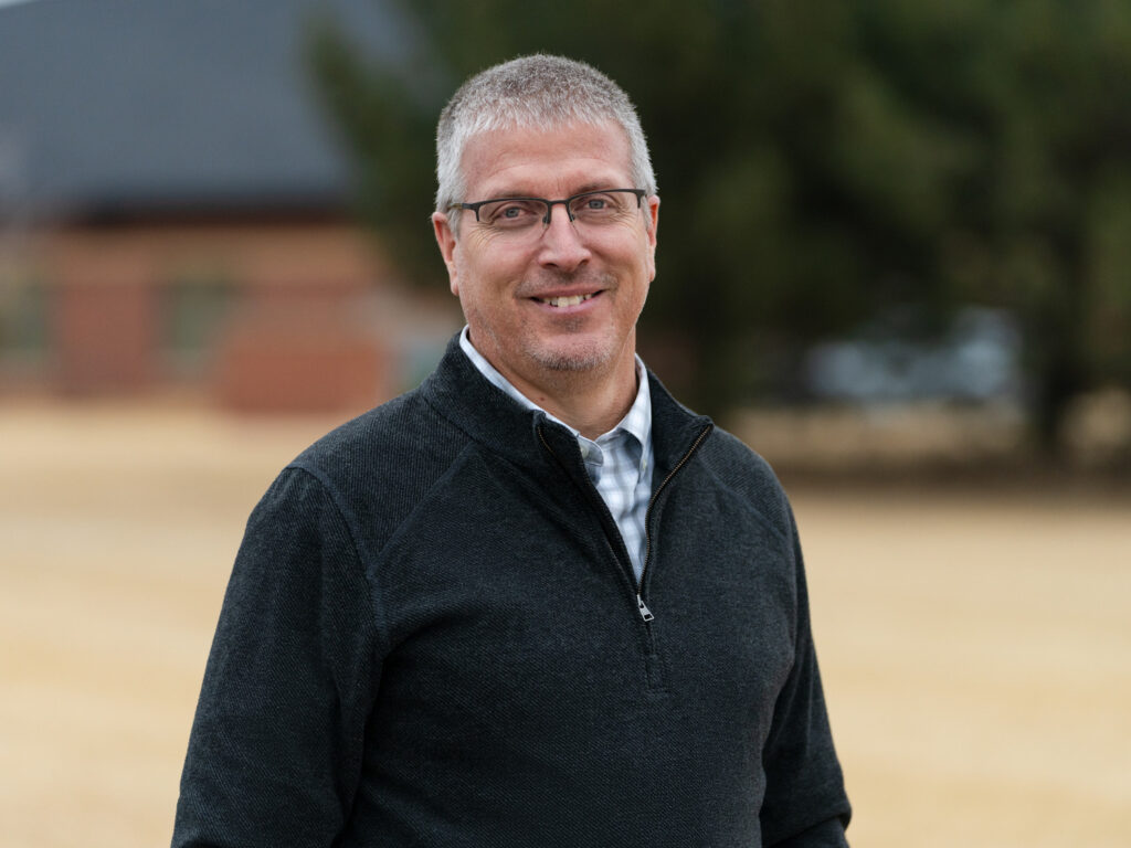 A man with short gray hair and glasses stands outdoors, wearing a dark zip-up sweater over a button-down shirt. He smiles at the camera. The background is blurred and shows trees and a building.