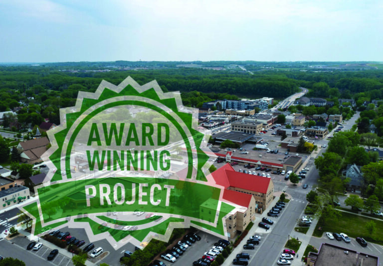 Aerial view of a small town with trees, roads, and buildings. Overlaid is a green and white badge with the words AWARD WINNING PROJECT in bold capital letters.