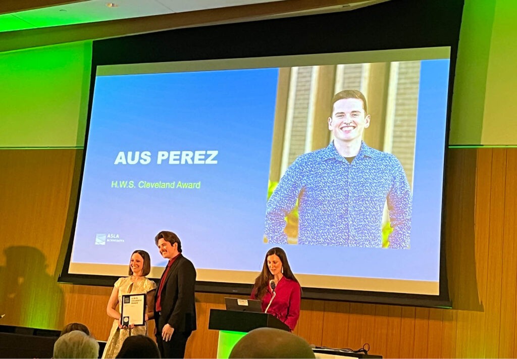 A man receives the Award on stage with two women. Behind them, a large screen shows a photo of a smiling man and the text Aus Perez, H.W.S. Cleveland Award.