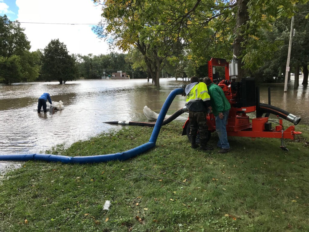 The image depicts a scene of flooding in a grassy area with several trees. Two individuals are working with a large red pump machine connected to blue hoses.